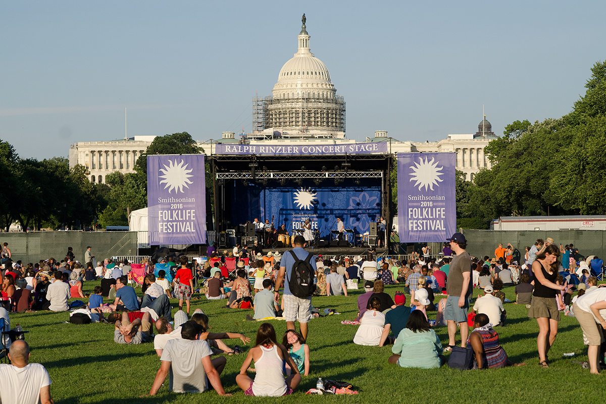 A crowd of people sit in the grass in front of an outdoor stage, flanked by purple banners for Smithsonian Folklife Festival. Behind it is the U.S. Capitol Building.