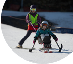 Ancfna and Leah work together as Leah skis down a groomed tail with modified skis on a sunny winter day.