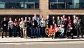 27 people lined up against a wall in bright sunshine.