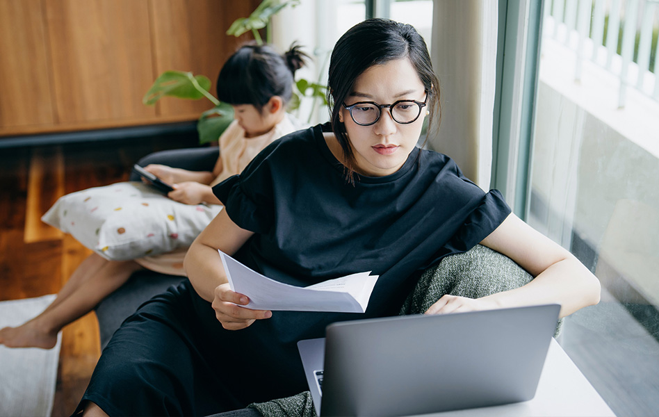 A woman sitting in front of a window holds a paper in one hand while she looks at a laptop.