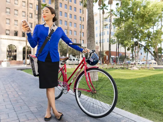 Woman in a blue cardigan and pencil skirt stands by her red bike, checking her phone, in downtown Tucson