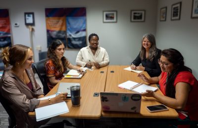 A group of women sit around a conference table, looking at a computer screen at one end.