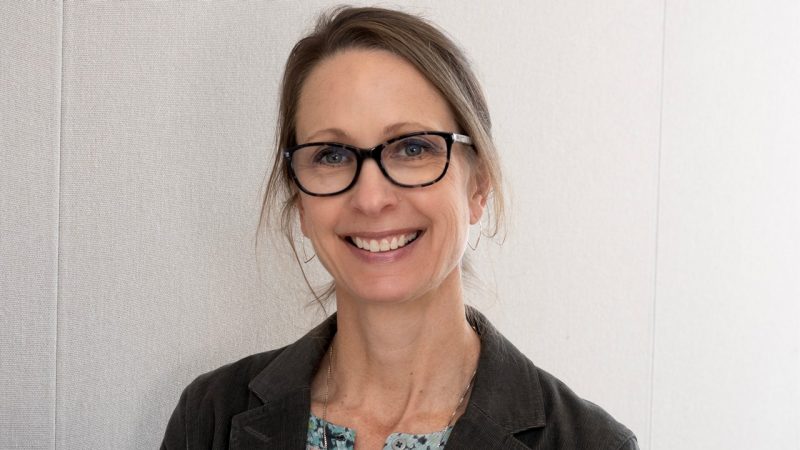 Headshot of Kelly standing against a white background. Her hair is pulled back and she wears tortoise shell-rimmed glasses.