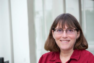 A woman wearing a red shirt and glasses stands in front of a window and smiles. 