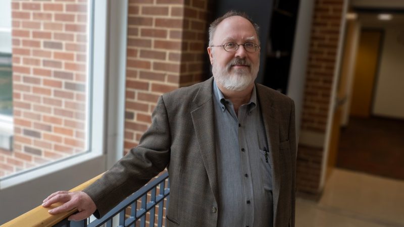 Mid-length portrait of a man with a gray beard, wearing glasses, standing between two brick columns.