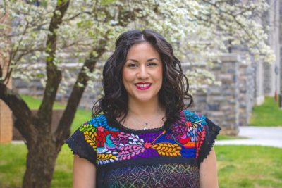 A woman with long, dark hair ,wearing a black shirt with colorful patterns, stands in front of a dogwood tree. She smiles into the camera. 