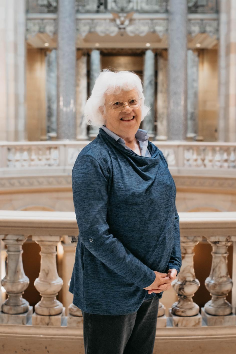 Peggy is wearing a gray sweater and smiling. She is inside the Capitol with the Rotunda behind her.