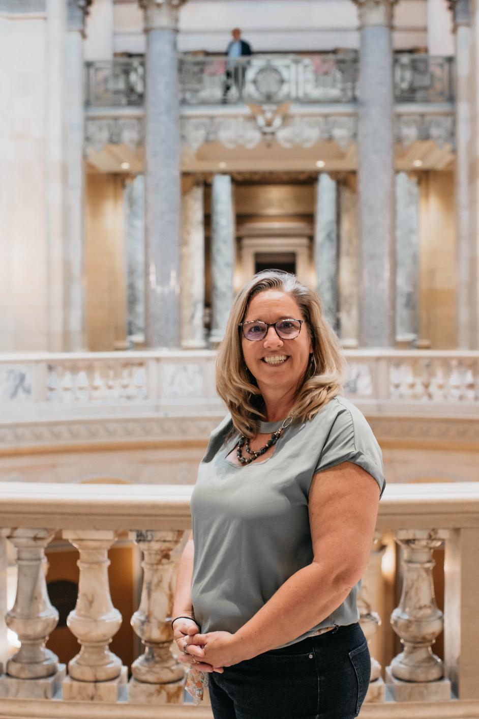 Brenda is wearing a gray business shirt and smiling. She is inside the Capitol with the Rotunda behind her.