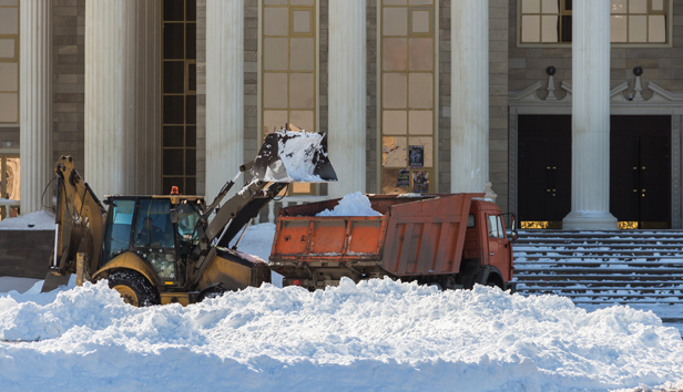 A backhoe shovels snow from in front of a government building into a dump truck