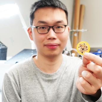 Scientist looking at the camera and holding up a small circular device slightly larger than a quarter