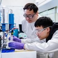 Two scientists in white lab coats in a lab peering together at equipment on a lab bench