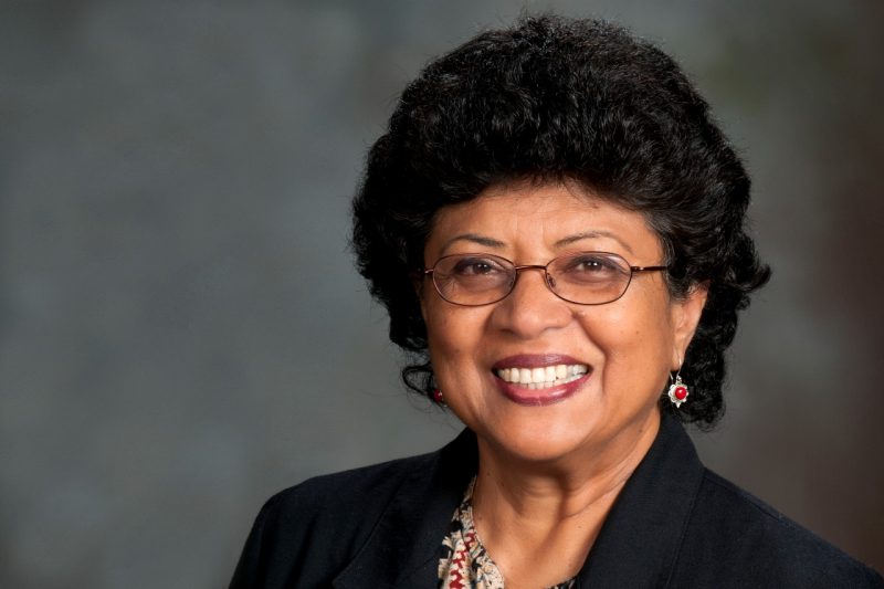 A woman, wearing a blazer and glasses, smiles for a headshot photo in front of a grey background. 