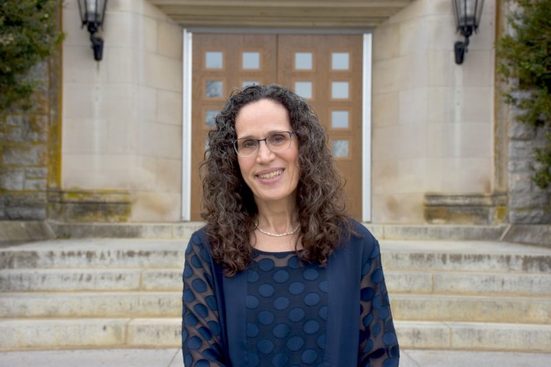 A woman with long black hair and glasses stand in front of the doors of Williams Hall.