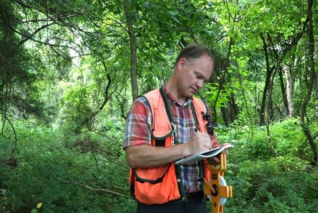 A person writes notes on a notebook while standing in a forest. 