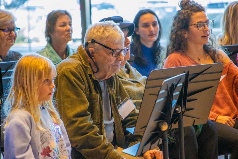 Members of a choir are seated in front of windows, and read lyrics as they practice.