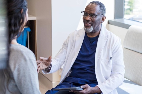 A physician sitting in a medical examination room holding a computer tablet and talking to a patient.