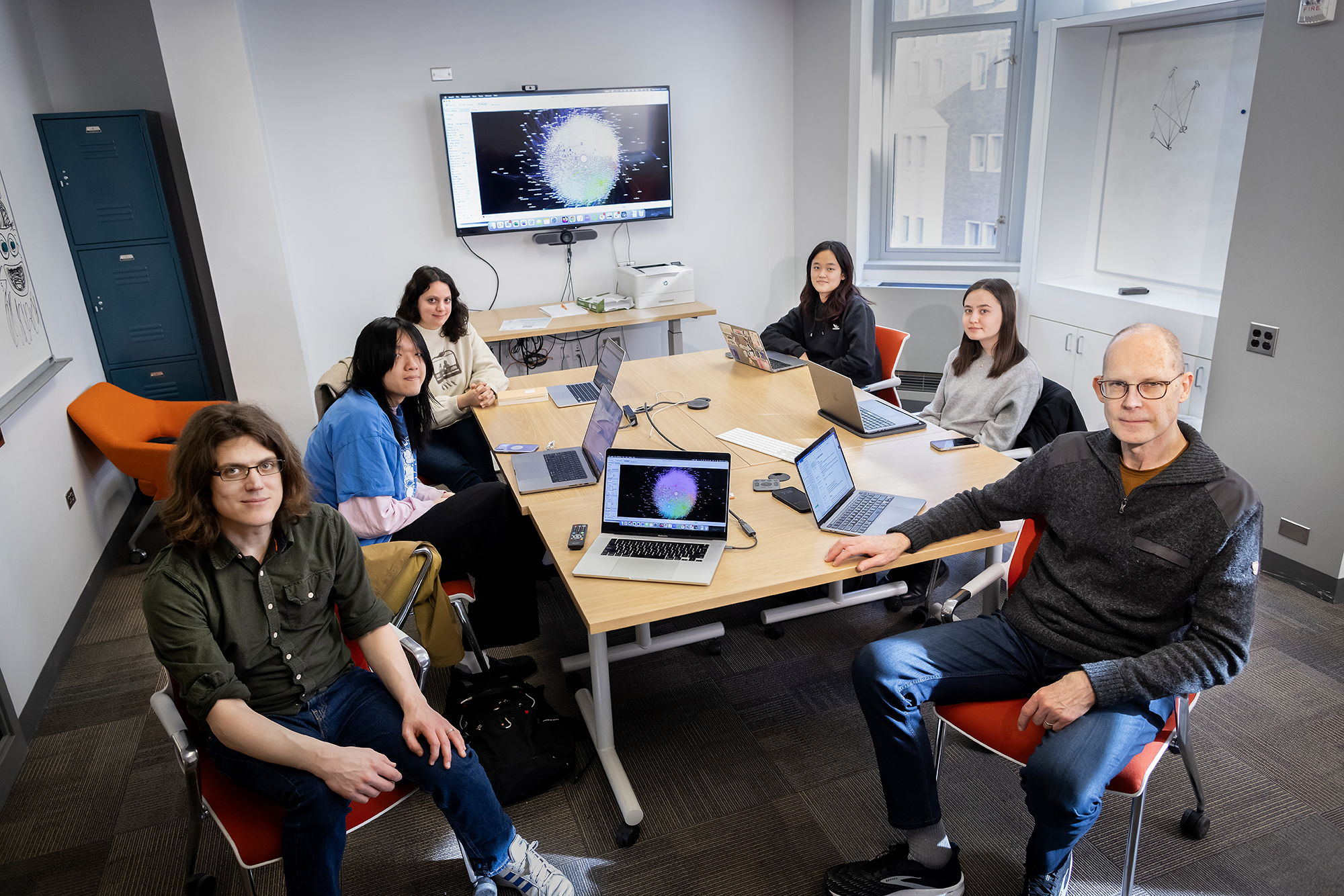 six people sitting at a table with laptop computers in front of them and a screen on the wall behind them