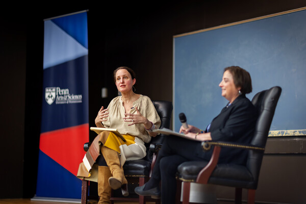 Sophia Rosenfeld and Beth Wenger sit on chairs on a wooden stage in front of a black board and a sign reading Penn Arts & Sciences, as Rosenfeld addresses the audience.