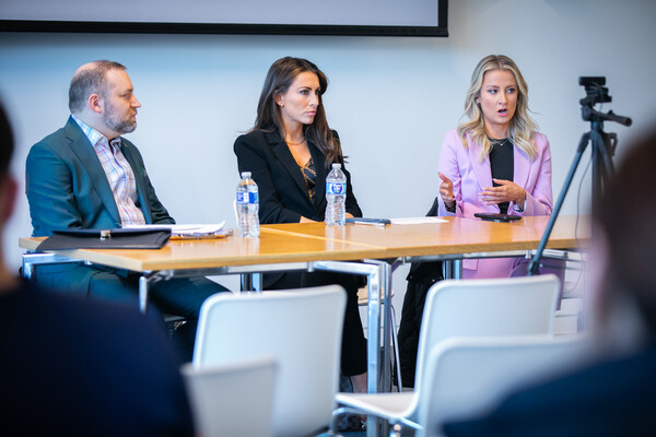 Three people sit at a table in front of a small video camera on a tripod as the person on the right gestures with her hands.