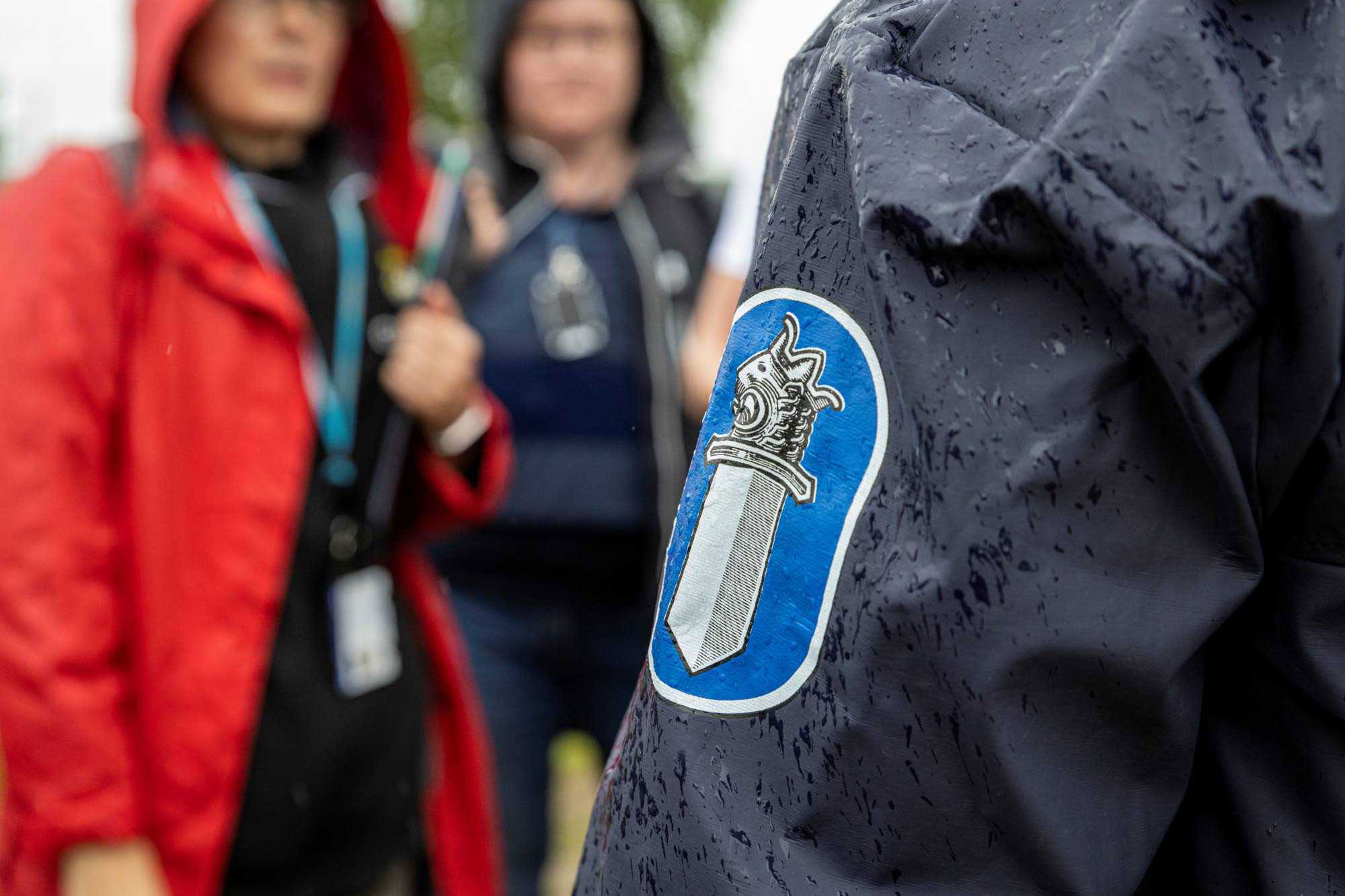 Kaksi poliisia ja pelastusviranomaisen edustaja keskustelevat. Etualalla poliisin sadetakin selkämys. 