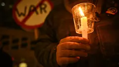 Close-up of a person holding a candle. Behind him is a sign with "War" crossed out