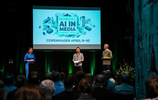 A photograph of the three conference organisers standing on a stage in front of a screen showing the conference's logo.