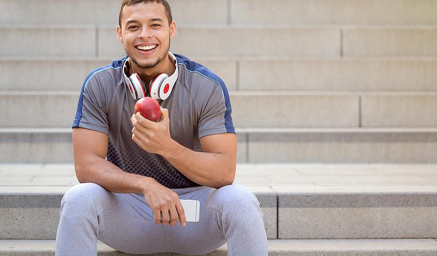 Un joven sonriente vestido con ropa deportiva sentado en un escalón comiendo una manzana