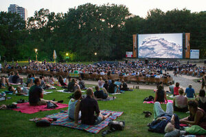 Zu sehen sind Menschen, die in einem Park am Boden sitzen und einer Leinwand zugewandt sind.