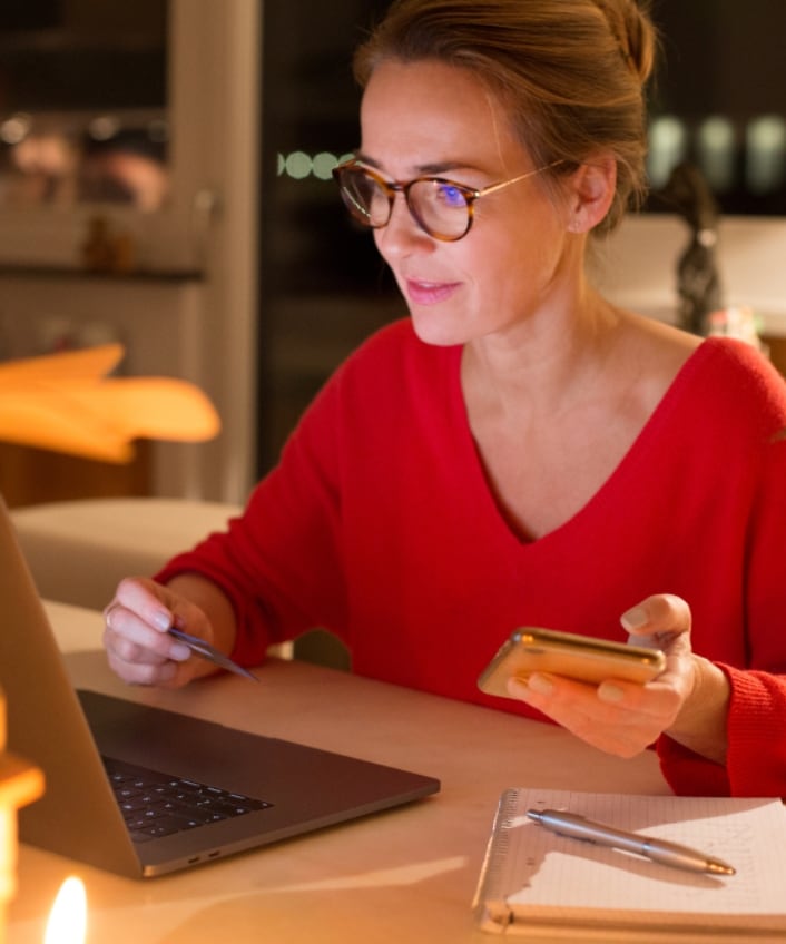 A woman in a red sweater sits at her laptop with her phone in one hand and her credit card in the other.