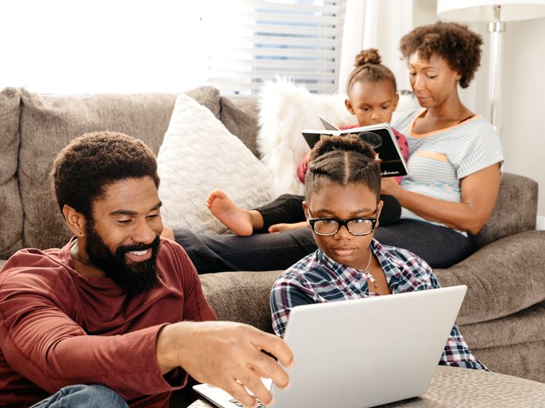 Family relaxing in living room, with mom reading to her young daughter on the sofa, and dad looking at a laptop resting on a coffee table with an older daughter