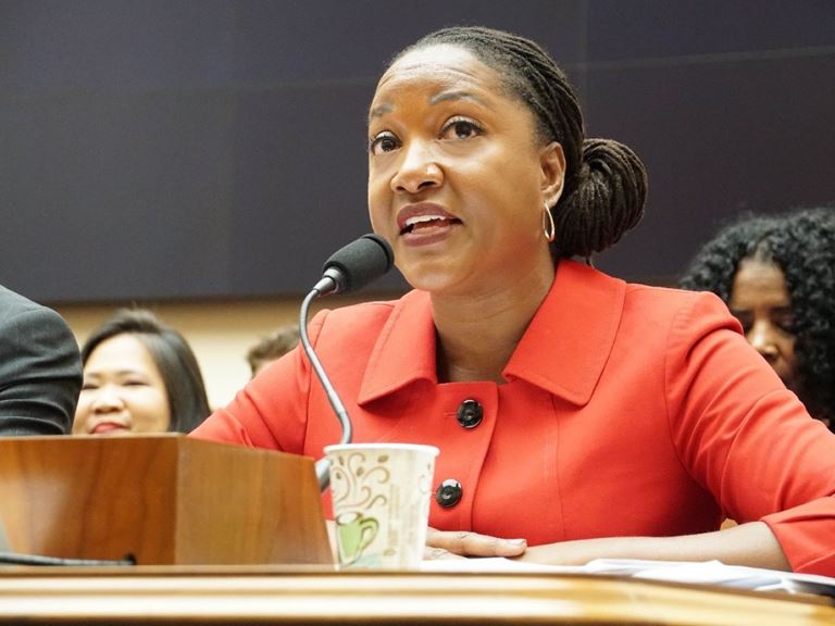 Seated woman with a red suit jacket speaking into a microphone at a public hearing