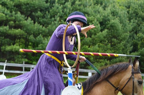 "Alanna on a light brown horse with black mane, throwing a spear through a ring"