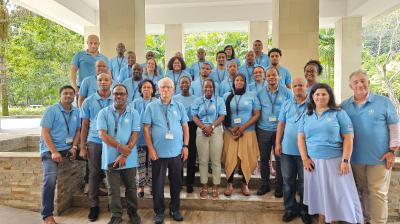 A group of 31 people wearing light blue polo shirts and name badges pose for a photo in a spacious indoor area with columns and greenery visible in the background.