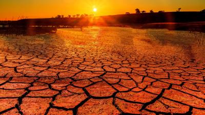 A cracked, dry landscape is illuminated by the setting sun. The sky is orange, and the silhouette of trees is visible in the background.