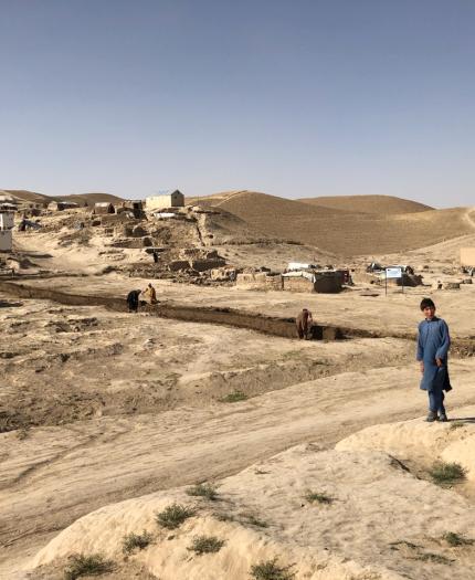 A man and a child walk on a dirt path in a rural, arid landscape with simple housing structures and people working in the distance.