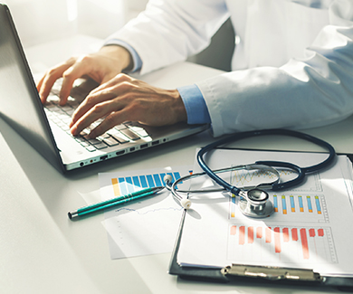 A physician's hands work at a laptop keyboard; a stethoscope and chart sit to one side of the computerc 