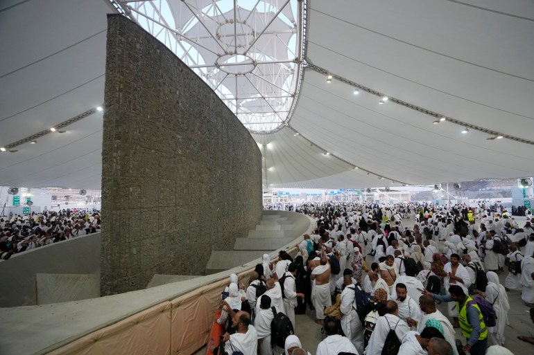 Pilgrims dressed in white gather under a sunshade to throw rocks at a big concrete pillar, one of the jamarat