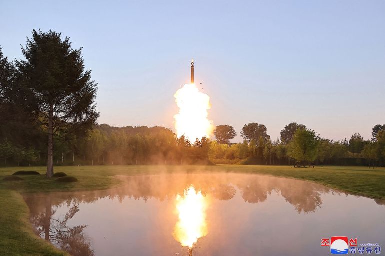 A missile taking off from an unconfirmed location in North Korea. There is a lake in front and manicured lawns fringed by trees. The missile is in the distance amid clouds of flame and white smoke. It is mainly black but has a section of black and white checks and a white nose cone.