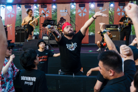 Southern Riot's lead singer Rudi on stage. He's wearing a black T-shirt and a red bandana around his head. The other band members are behind. There are crowds around him. He is holding the microphone out to the fans