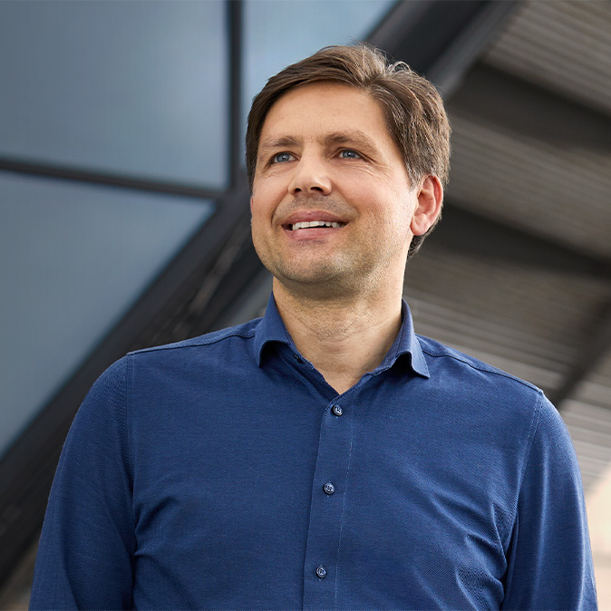 Greg standing outdoors in front of a window-paned wall, looking to the left, smiling.