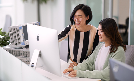 Ella standing beside a colleague who is seated in front of an Apple display.