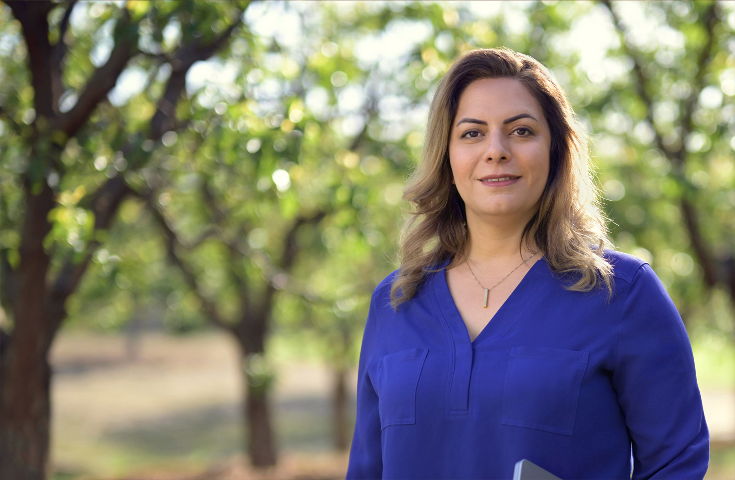 Desireh sorri para a câmera. Ela está em pé e segura um MacBook. 