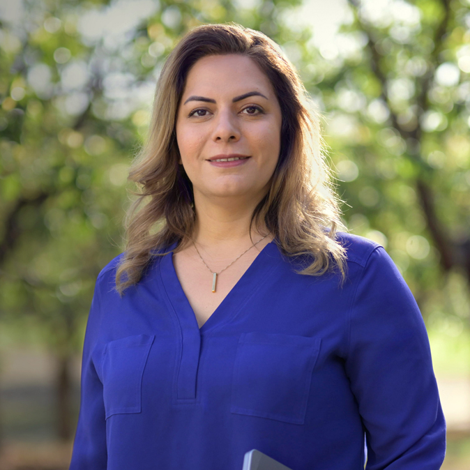 Desireh sorri para a câmera. Ela está em pé e segura um MacBook. 