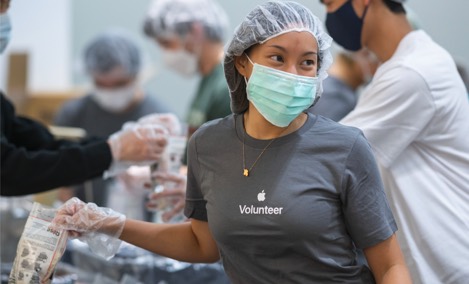 Estagiária da Apple, vestindo uma camiseta de voluntário da Apple, sorri e olha para o lado enquanto embala itens em um evento de voluntariado.