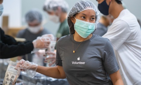 Een stagiair van Apple in een t-shirt voor Apple vrijwilligers lacht en kijkt opzij bij het inpakken van artikelen tijdens een vrijwilligersevenement.