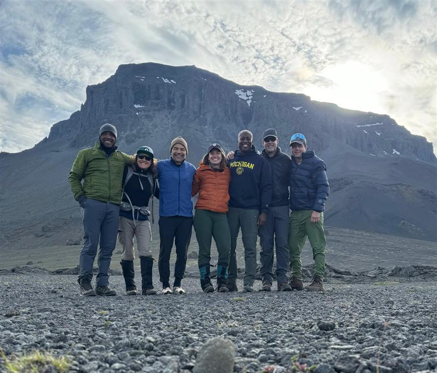 Seven people pose on a rocky terrain in front of a mountain.