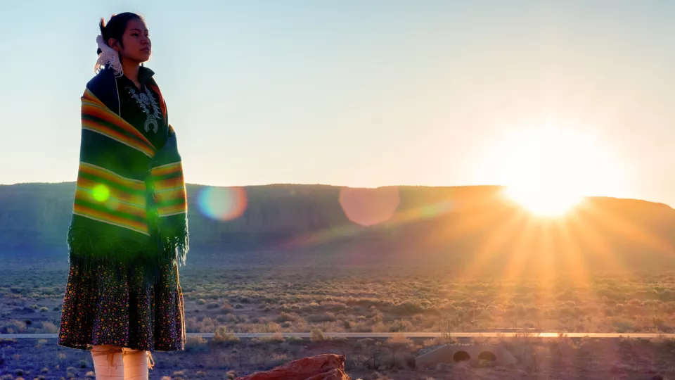 A woman wearing a skirt and moccasins wrapped in a shawl looks out on a mountainous landscape.