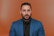 Josh Altman wearing a blue jacket in front of an orange backdrop