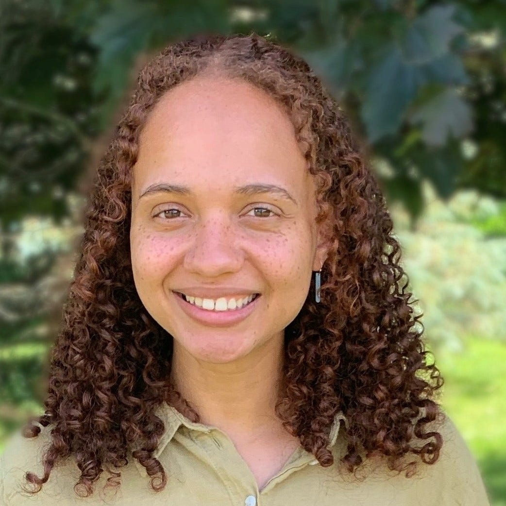 young woman in front of tree with curly reddish/brown hair and freckles, beige top with collar 