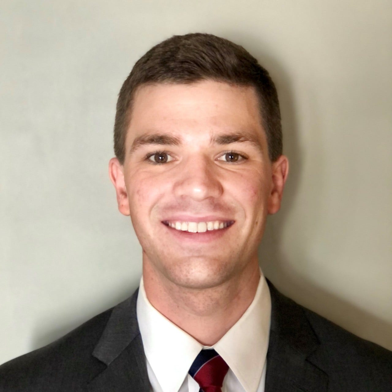 white man against beige wall wearing dark gray jacket, white shirt, and red and blue tie